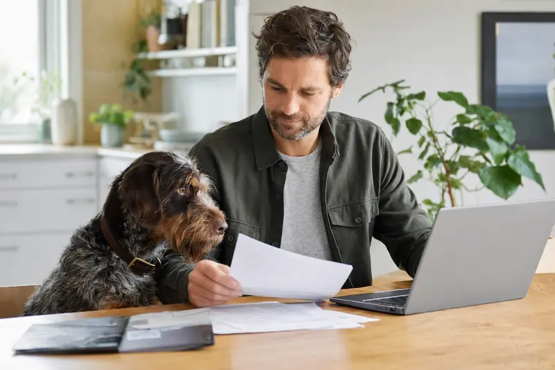 Ein Gammel Dansk Honsehund sitzt ruhig neben seinem Besitzer am Küchentisch, auf dem Versicherungsunterlagen und ein Laptop liegen.