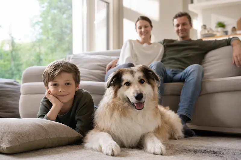 Ein freundlicher Hund liegt entspannt neben einem Kind im hellen Wohnzimmer, während die Eltern im Hintergrund eine sichere Familienatmosphäre schaffen.
