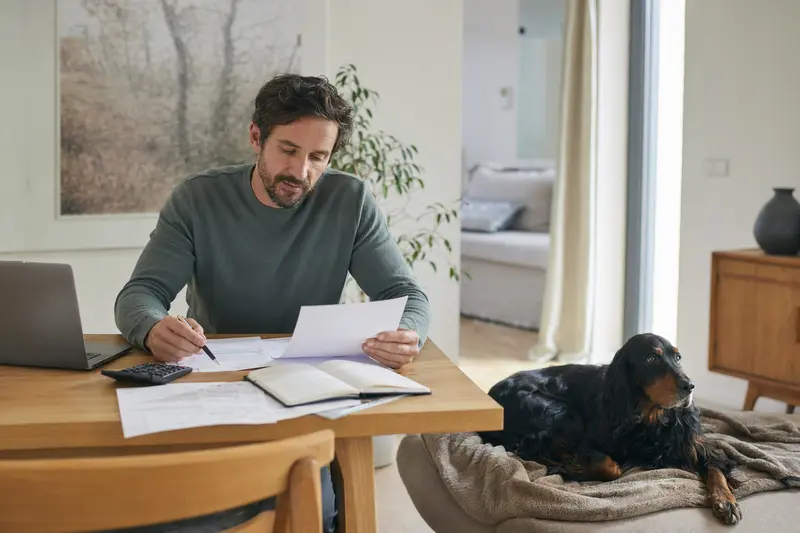Ein Hundehalter sitzt mit einer Tierarztrechnung am Tisch, während sein entspannter Gordon Setter auf einer Decke liegt und eine Hundekrankenversicherung als Lösung des Tierarztkosten-Themas nahelegt.