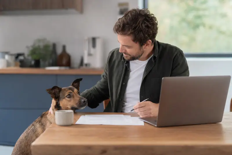Ein Gos Rater Valencia sitzt ruhig neben seinem Besitzer am Küchentisch, auf dem Versicherungsunterlagen und ein Laptop mit Vergleichsseite liegen, passend zum Thema Hundekrankenversicherung.