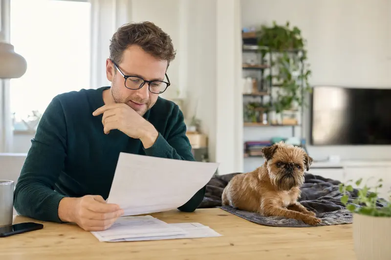 Ein Hundebesitzer sitzt mit einer Tierarztrechnung am Tisch, während ein entspannter Griffon Bruxellois auf einer Decke liegt.