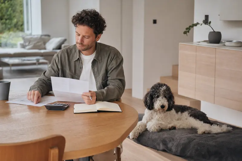 Ein Hundehalter sitzt mit einer Tierarztrechnung am Tisch, während ein entspannter Großpudel auf einer Decke daneben liegt – ein ruhiger Moment zum Thema Hundekrankenversicherung und Tierarztkosten.