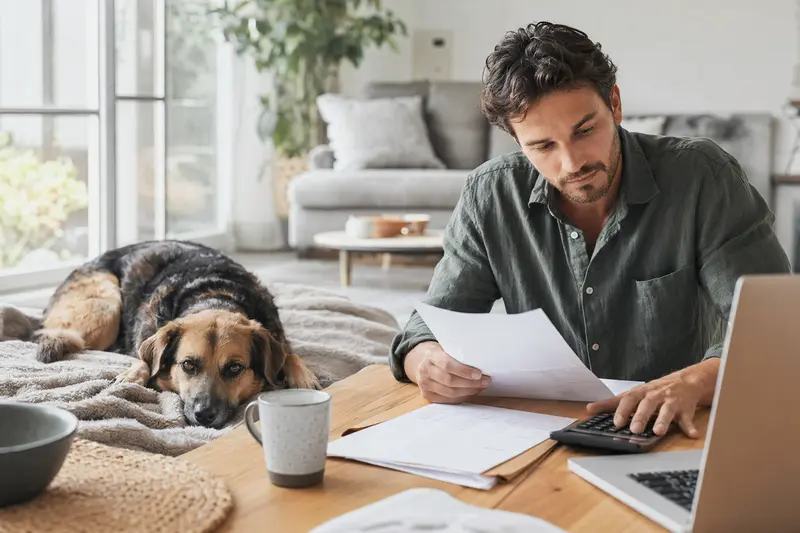 Ein Hundehalter sitzt mit einer Tierarztrechnung am Tisch, während ein entspannter Haldenlaufhund auf einer Decke in der hellen Wohnung liegt – ein ruhiger Moment rund um Hundekosten und Versicherung.