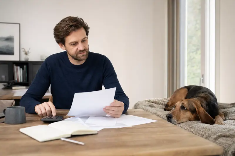 Ein Hundebesitzer sitzt mit einer Tierarztrechnung am Tisch, während ein entspannter Haldenstövare auf einer Decke daneben liegt, passend zum Thema Hundekrankenversicherung.