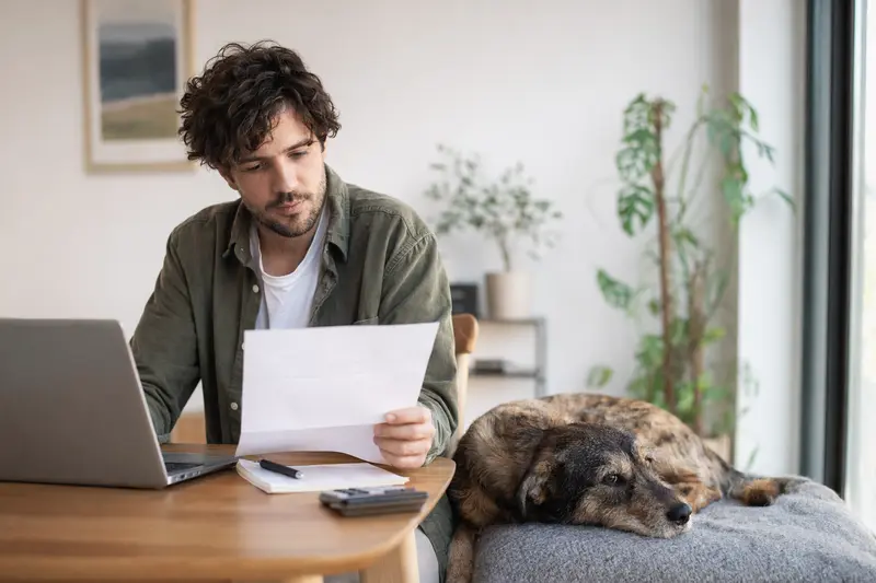 Ein Hundehalter sitzt mit einer Tierarztrechnung am Tisch, während ein entspannter Haldenstoever-Hund auf einer Decke liegt und das Thema Hundekrankenversicherung ruhig veranschaulicht.