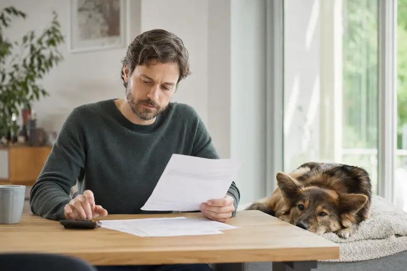 Ein Hundehalter sitzt mit einer Tierarztrechnung am Tisch, während ein entspannter Harzer Fuchs auf einer Decke im hellen Wohnzimmer liegt und die Situation ruhig wirkt.