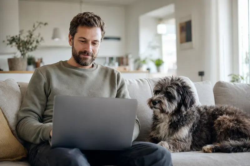 Ein Hundebesitzer mit einem Hawaiian Poi Dog sitzt mit Laptop auf dem Sofa und vergleicht Versicherungen.