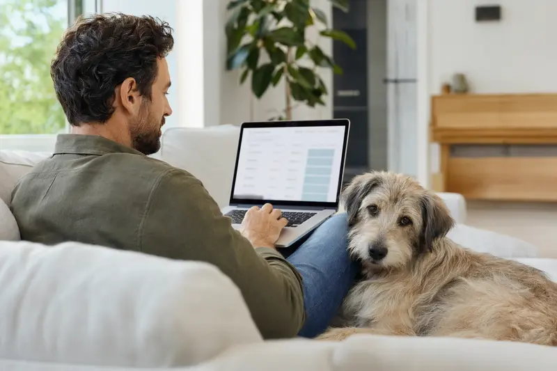 Ein Hundebesitzer arbeitet am Laptop auf dem Sofa, während ein Hellenikos Ichnilatis Hund neben ihm sitzt, was eine sichere und informierte Entscheidung über die Tierversicherung symbolisiert.
