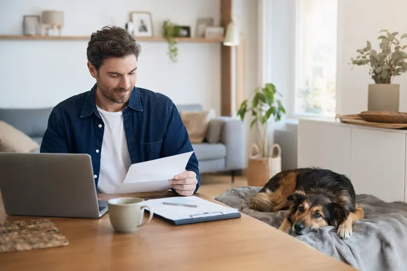 Ein Hundebesitzer sitzt mit einer Tierarztrechnung am Tisch, während ein entspannter Herder Hund auf einer Decke liegt und das Thema Tierarztkosten bei der Hundever­sicherung ruhig vermittelt.
