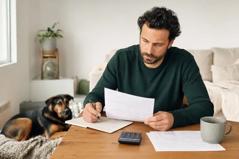 Ein Hundebesitzer sitzt mit einer Tierarztrechnung am Tisch, während ein ruhiger Huntaway auf einer Decke in der hellen Wohnung liegt und die Situation gelassen wirkt.
