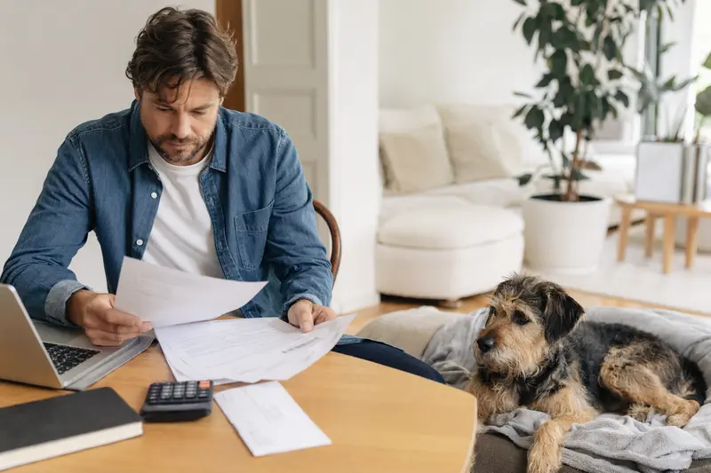 Ein Hundehalter sitzt mit einer Tierarztrechnung am Tisch, während sein entspannter Hybridhund auf einer Decke in der hellen Wohnung liegt und das Thema Hundekrankenversicherung ruhig einordnet.