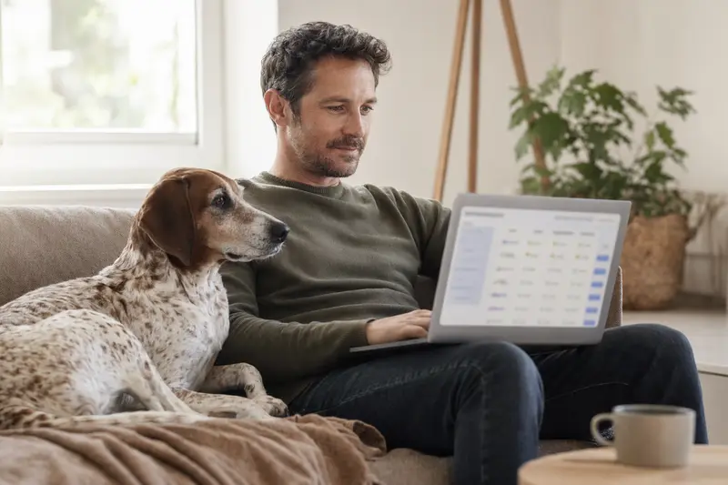 Ein Hundebesitzer sitzt mit einem Laptop auf dem Sofa, während ein Hygienbracke neben ihm liegt und ein Versicherungsvergleich auf dem Bildschirm angezeigt wird.