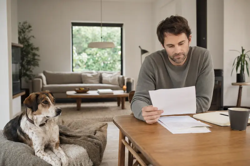 Ein Hundebesitzer sitzt mit Tierarztrechnung am Tisch, während ein entspannter Hygenlaufhund auf einer Decke in der hellen Wohnung liegt und das Thema Hundeversicherung ruhig vermittelt.
