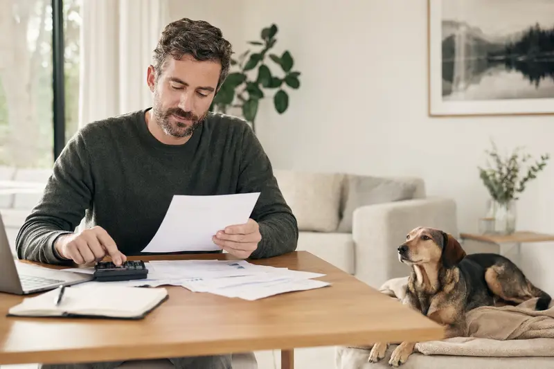 Besitzer mit Tierarztrechnung am Tisch, daneben liegt ein entspannter Illyrischer Laufhund auf einer Decke in einer hellen Wohnung, passend zum Thema Hundekrankenversicherung.