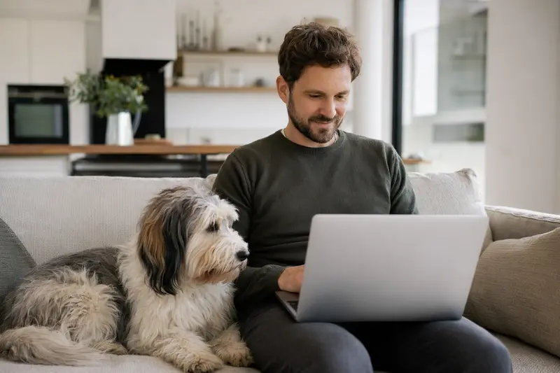 Ein Hundebesitzer arbeitet auf dem Sofa an seinem Laptop, während ein Indian Beardog neben ihm sitzt, was eine sichere und informierte Entscheidung über Tierversicherungen suggeriert.