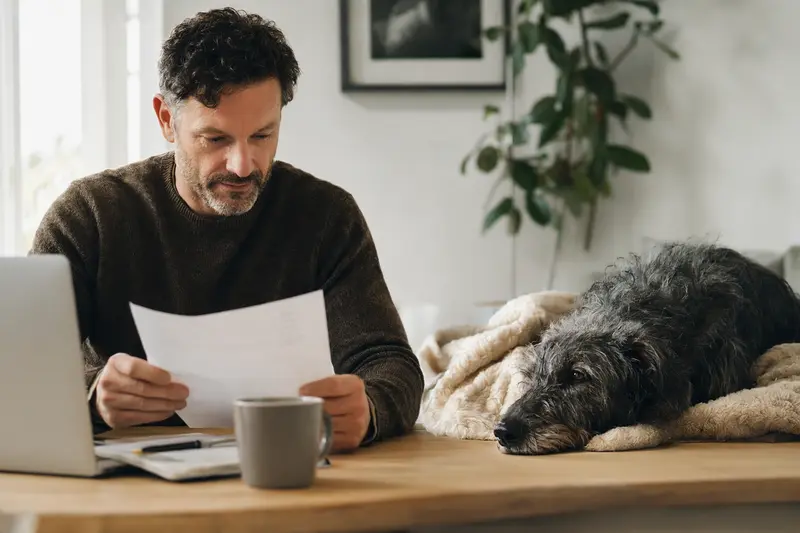 Besitzer sitzt mit Tierarztrechnung am Tisch, während ein entspannter Irischer Wolfshund auf einer Decke daneben liegt – ein ruhiges Motiv zum Thema Hundekrankenversicherung.