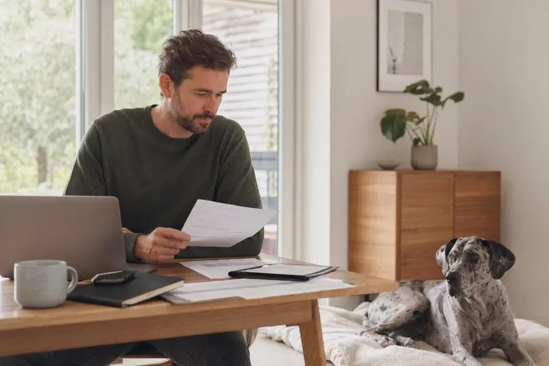 Ein Hundebesitzer sitzt mit einer Tierarztrechnung am Tisch, während seine Italienische Dogge entspannt auf einer Decke neben ihm liegt – ein ruhiges Motiv rund um Tierarztkosten und Hundesicherung.