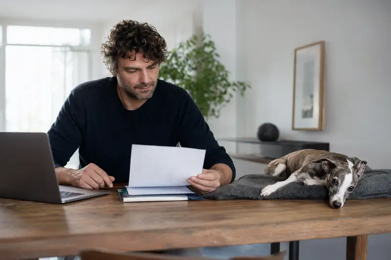 Ein Hundehalter sitzt mit einer Tierarztrechnung am Tisch, während ein entspannter Italienisches Windspiel Hund ruhig auf einer Decke liegt, passend zum Thema Hundekrankenversicherung.