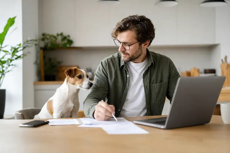 Ein Jack Russell Terrier sitzt ruhig neben seinem Besitzer am Küchentisch, während Versicherungsunterlagen und ein Laptop liegen.