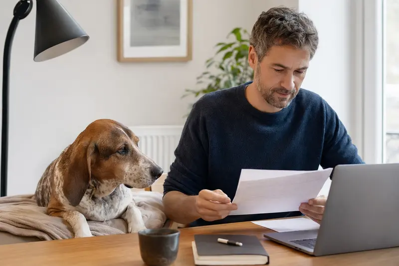 Ein Besitzer sitzt mit einer Tierarztrechnung am Tisch, während ein entspannter Jura Laufhund auf einer Decke daneben liegt – ein ruhiges Motiv zum Thema Hundekrankenversicherung.