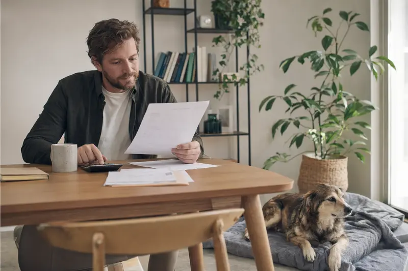 Ein Hundebesitzer sitzt mit einer Tierarztrechnung am Tisch, während sein ruhiger Kars-Hund entspannt auf einer Decke liegt.