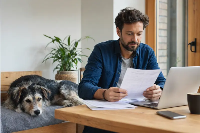 Ein Hundehalter sitzt mit Tierarztrechnung am Tisch, während ein entspannter Kaikadi Hund auf einer Decke liegt – ein ruhiges Bild zum Thema Hundeversicherung und Tierarztkosten.