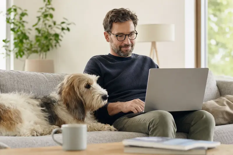 Besitzer mit Laptop auf dem Sofa und kleinem Basset Griffon Vendéen Hund in einer hellen Wohnung, konzentriert auf den Versicherungsvergleich.