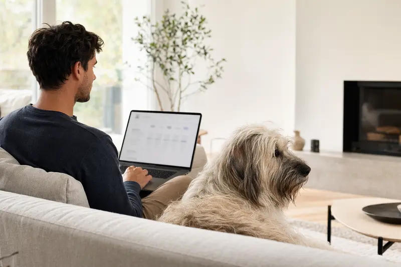 Ein Hundebesitzer sitzt auf dem Sofa mit einem Komondor neben sich und betrachtet Versicherungsoptionen auf seinem Laptop.