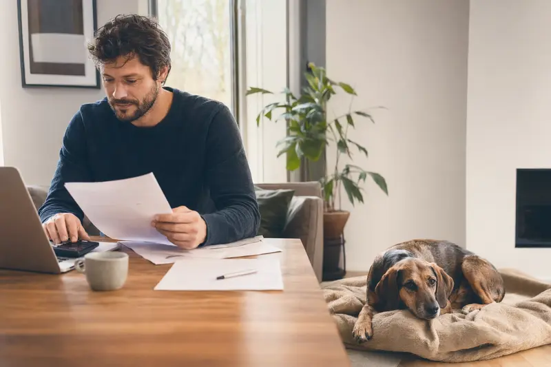 Ein Hundebesitzer sitzt mit einer Tierarztrechnung am Tisch, während ein entspannter Kopov Bracke Hund auf einer Decke daneben liegt.
