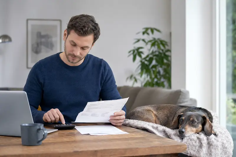 Ein Hundebesitzer sitzt mit einer Tierarztrechnung am Tisch, während ein entspannter Kurzhaardackel auf einer Decke daneben liegt – ein realistisches Bild zur Tierarztkosten-Absicherung für Hunde.