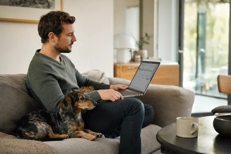 Ein Hundebesitzer mit einem Kurzhaardackel sitzt auf dem Sofa und vergleicht Tierversicherungen auf seinem Laptop in einer hellen Wohnung.