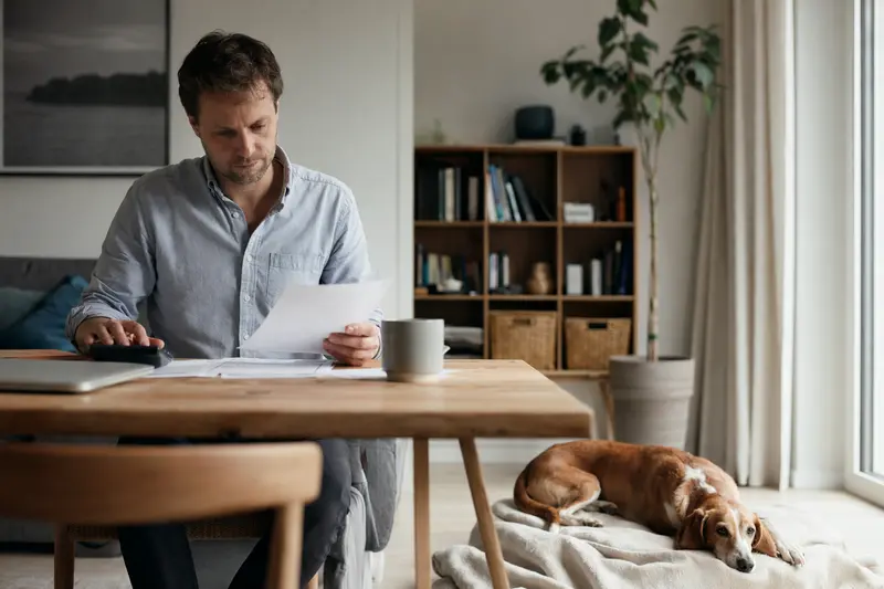 Besitzer mit Tierarztrechnung am Tisch, daneben liegt eine ruhige Litauische Bracke auf einer Decke in heller Wohnung, als Bild zu Tierarztkosten und Hundeversicherung.