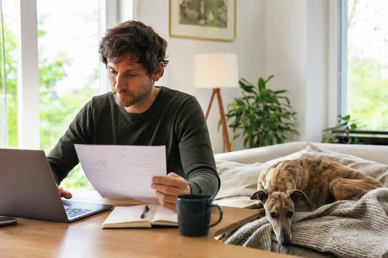 Ein Hundehalter sitzt mit einer Tierarztrechnung am Tisch, während ein entspannter Magyar Agar auf einer Decke neben ihm liegt.
