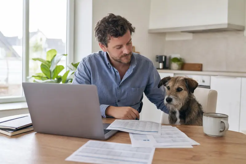 Ein ruhiger Mallorqui Hund sitzt neben seinem Besitzer am Küchentisch, wo Versicherungsunterlagen und ein Laptop liegen.