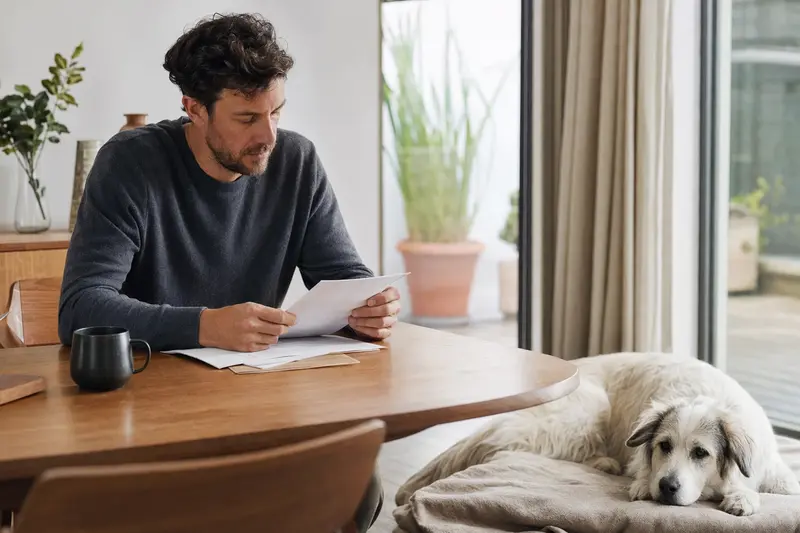 Ein Hundehalter sitzt mit einer Tierarztrechnung am Tisch, während ein entspannter Maremmaner Hirtenhund auf einer Decke daneben liegt und das Thema Tierarztkosten für Hunde ruhig veranschaulicht.