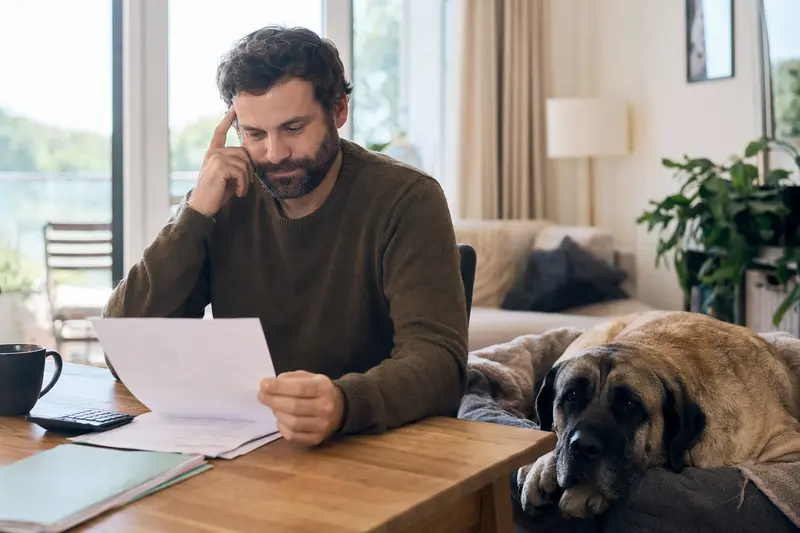 Ein Hundehalter sitzt mit einer Tierarztrechnung am Tisch, während ein entspannter Mastin extremeno auf einer Decke liegt und das Thema Tierarztkosten im Rahmen einer Hundekrankenversicherung ruhig dargestellt wird.