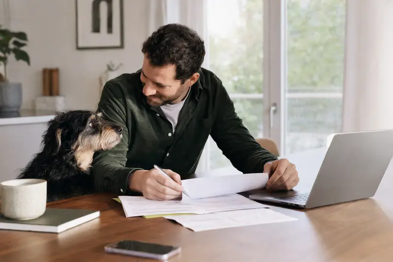 Ein Matin Hund sitzt ruhig neben seinem Besitzer am Küchentisch, auf dem Versicherungsunterlagen und ein Laptop liegen.