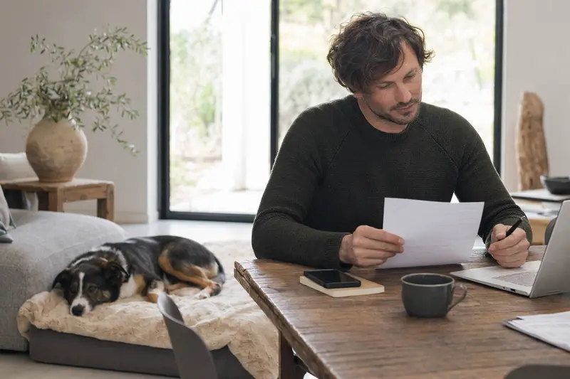 Ein Hundebesitzer sitzt mit einer Tierarztrechnung am Tisch, während ein entspannter McNab Hund ruhig auf einer Decke liegt und das Thema Hundekrankenversicherung im Alltag veranschaulicht.