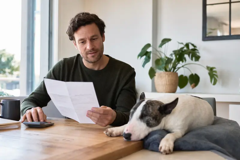 Ein Hundebesitzer sitzt mit einer Tierarztrechnung am Tisch, während ein entspannter Mini-Bullterrier auf einer Decke liegt und den Bezug zu Tierarztkosten und Hundekrankenversicherung zeigt.