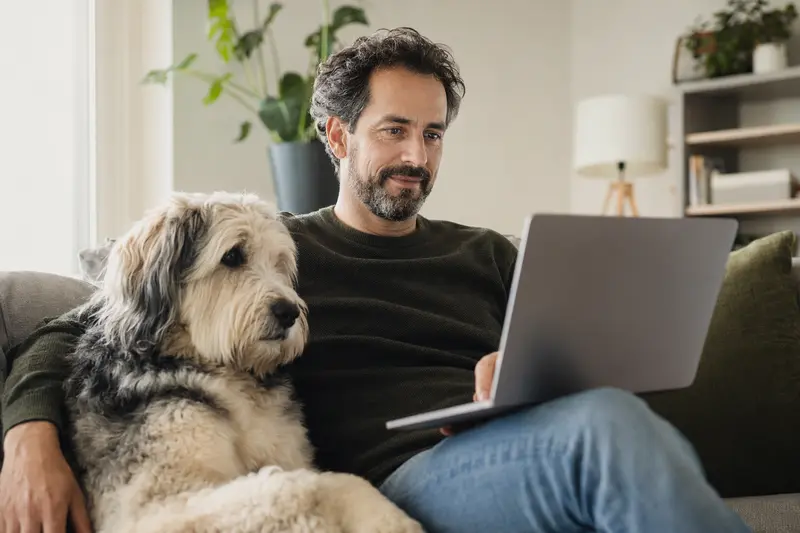Ein Hundebesitzer arbeitet entspannt auf dem Sofa mit seinem Mioritic-Hund, während er einen Versicherungsvergleich auf dem Laptop betrachtet.