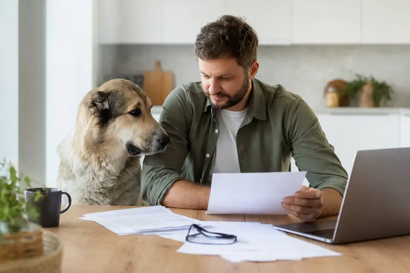 Ein Mittelasiatischer Schäferhund sitzt ruhig neben seinem Besitzer am Küchentisch mit Versicherungsunterlagen und einem Laptop zu einer Hundekrankenversicherung.