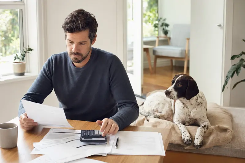 Ein Hundehalter sitzt mit einer Tierarztrechnung am Tisch, während ein entspannter Navarro Pointer auf einer Decke neben ihm liegt und die Tierarztkosten bei Hund und Versicherung ruhig veranschaulicht.