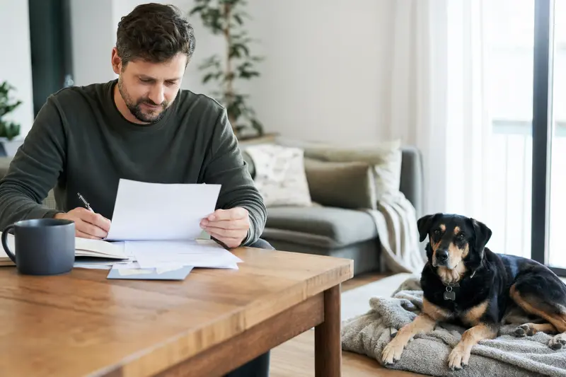 Ein Hundebesitzer sitzt mit einer Tierarztrechnung am Tisch, während ein entspannter Neuseeländer Huntaway auf einer Decke daneben liegt – ein ruhiger Moment mit Bezug zu Tierarztkosten und Hundeversicherung.