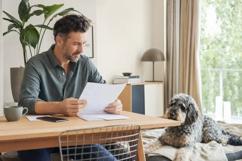Ein Hundebesitzer sitzt mit einer Tierarztrechnung am Tisch, während ein entspannter Niederländischer Schafspudel auf einer Decke neben ihm liegt.