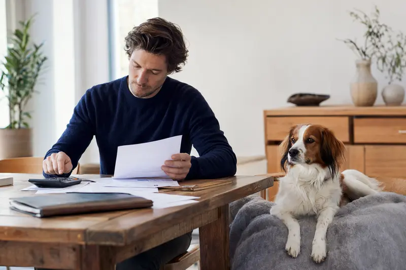Ein Hundebesitzer sitzt am Tisch und prüft eine Tierarztrechnung, während ein entspannter Niederländisches Kooikerhondje auf einer Decke daneben liegt.