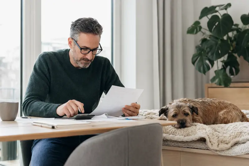 Ein Hundehalter sitzt mit einer Tierarztrechnung am Tisch, während ein entspannter Norfolk-Terrier auf einer Decke liegt und die Situation ruhig wirkt.