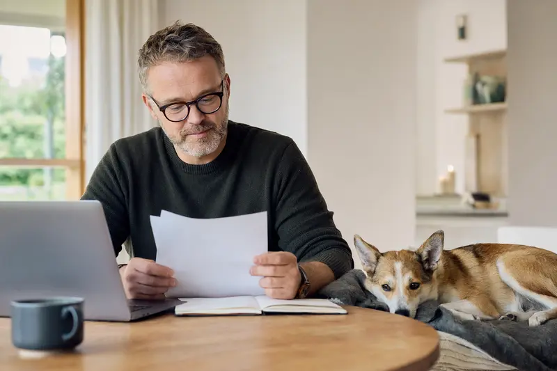 Ein Hundehalter sitzt mit einer Tierarztrechnung am Tisch, während sein entspannter Norwegischer Lundehund auf einer Decke liegt, passend zum Thema Hundekrankenversicherung.
