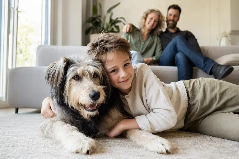 Ein freundlicher Hund liegt entspannt neben einem Kind im Wohnzimmer in einer sicheren Familienatmosphäre, während die Eltern im Hintergrund stehen.