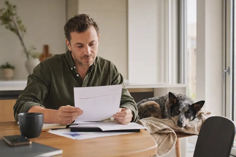 Ein Hund vom Typ Ormskirk Heeler liegt ruhig auf einer Decke, während sein Besitzer am Tisch eine Tierarztrechnung prüft und eine Hundekrankenversicherung im Blick behält.