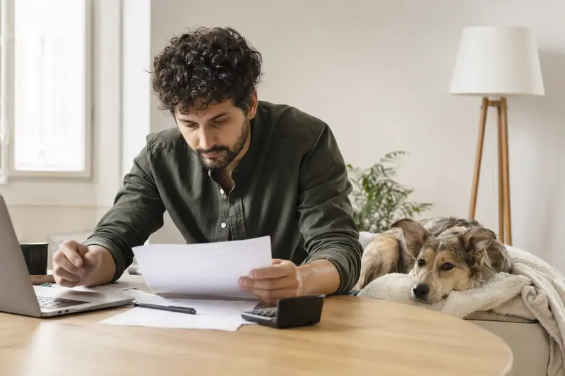Ein Hundebesitzer sitzt mit einer Tierarztrechnung am Tisch, während ein entspannter Pai Hund auf einer Decke liegt und das Thema Tierarztkosten für Hunde ruhig wirkt.